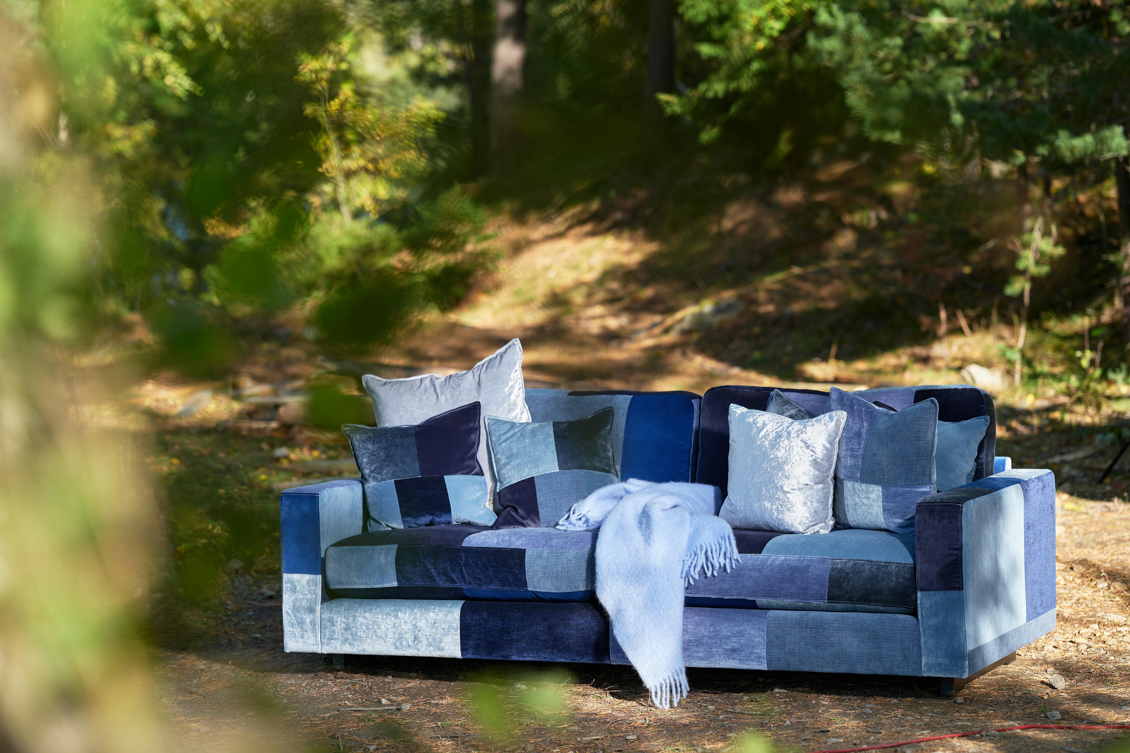 Valen 224 soffa i blått Sweef patchtyg. Soffan står i en skog, i soffan finns kuddar i olika storlekar och en blå filt.