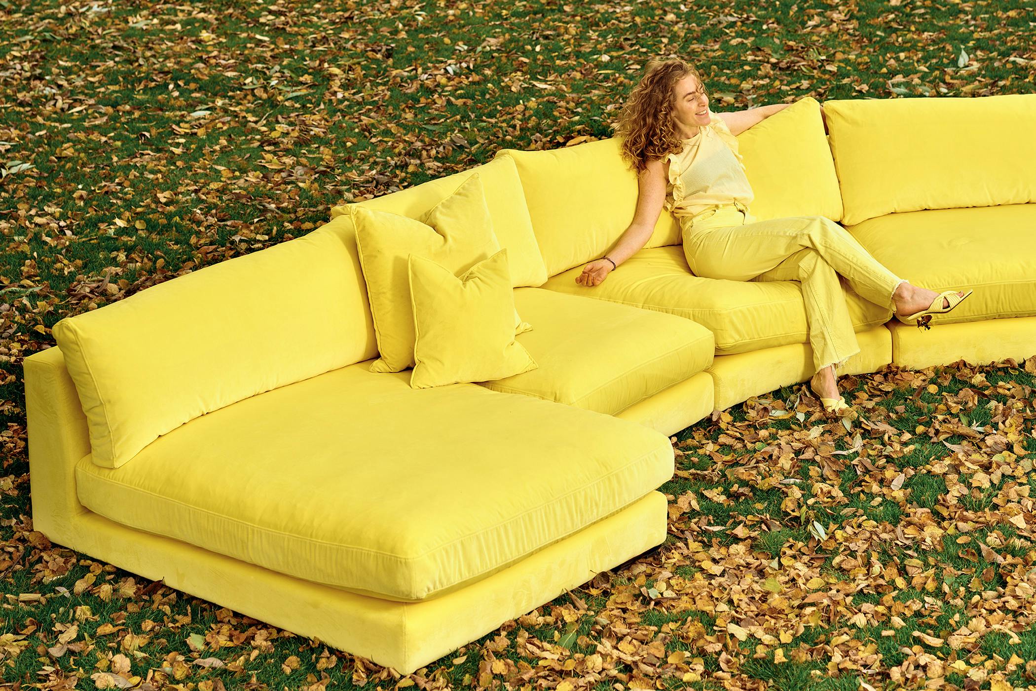 Svängd modulsoffa i gul polyestersammet med djupa sittkuddar, låga ryggkuddar och med låg sitthöjd.