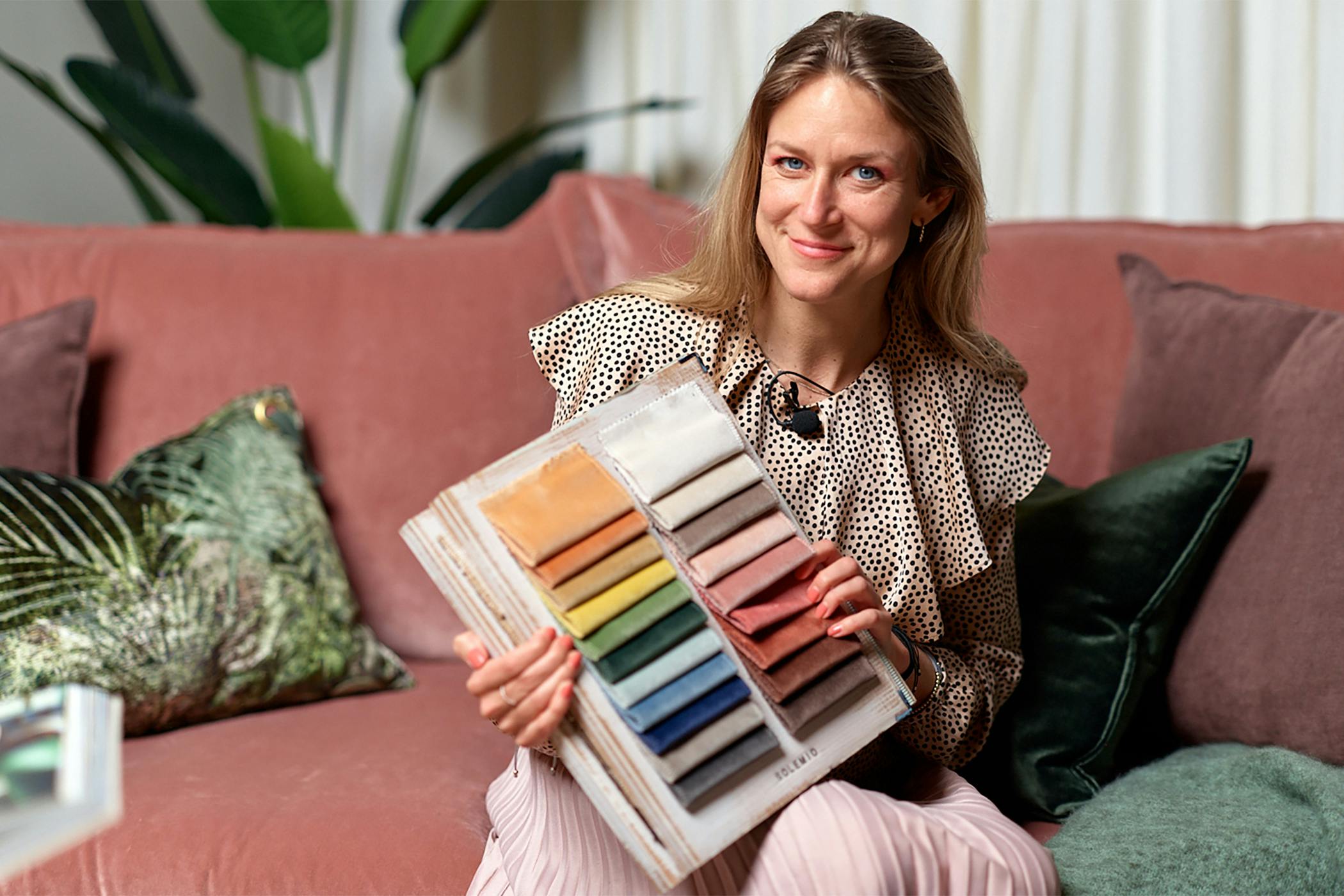 En professionell färgforskare, Sara Garanty, sitter bekvämt i en modern rosa soffa och presenterar en tygkarta med ett urval av färger för anpassningsbara soffklädsel på Sweef.