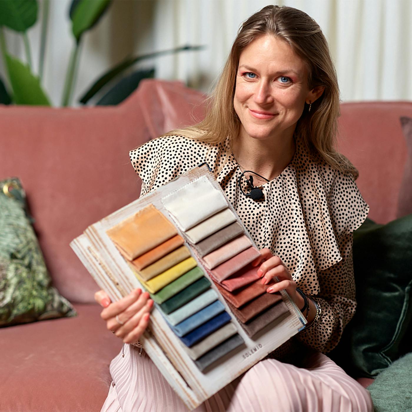 En professionell färgforskare, Sara Garanty, sitter bekvämt i en modern rosa soffa och presenterar en tygkarta med ett urval av färger för anpassningsbara soffklädsel på Sweef. Hennes klädsel och smycken är elegant samordnade med de mjuka tonerna i rummet, som illustrerar konceptet av färgpsykologi i heminredning. Denna bild tjänar som en visuell hjälp för att betona hur valet av soffans färg kan påverka en persons känslor och atmosfären i ett rum.
