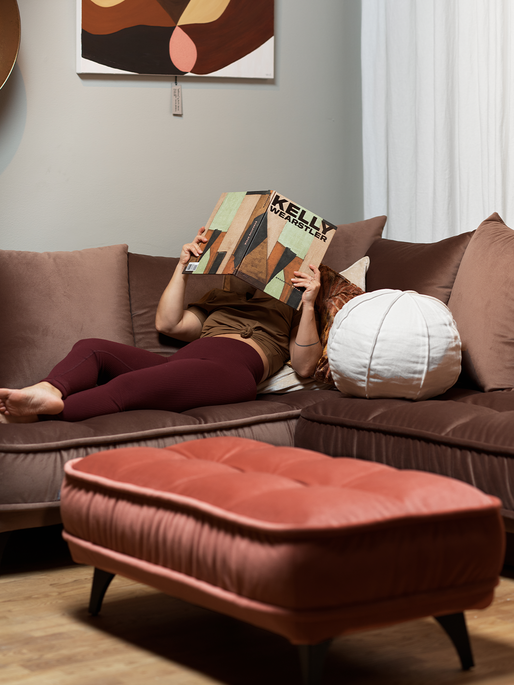 Brun bäddsoffa Vargen i sammet. En kvinna ligger i soffan och läser en tidning, bredvid henne är en bollkudde. Framför soffan står en fotpall. 